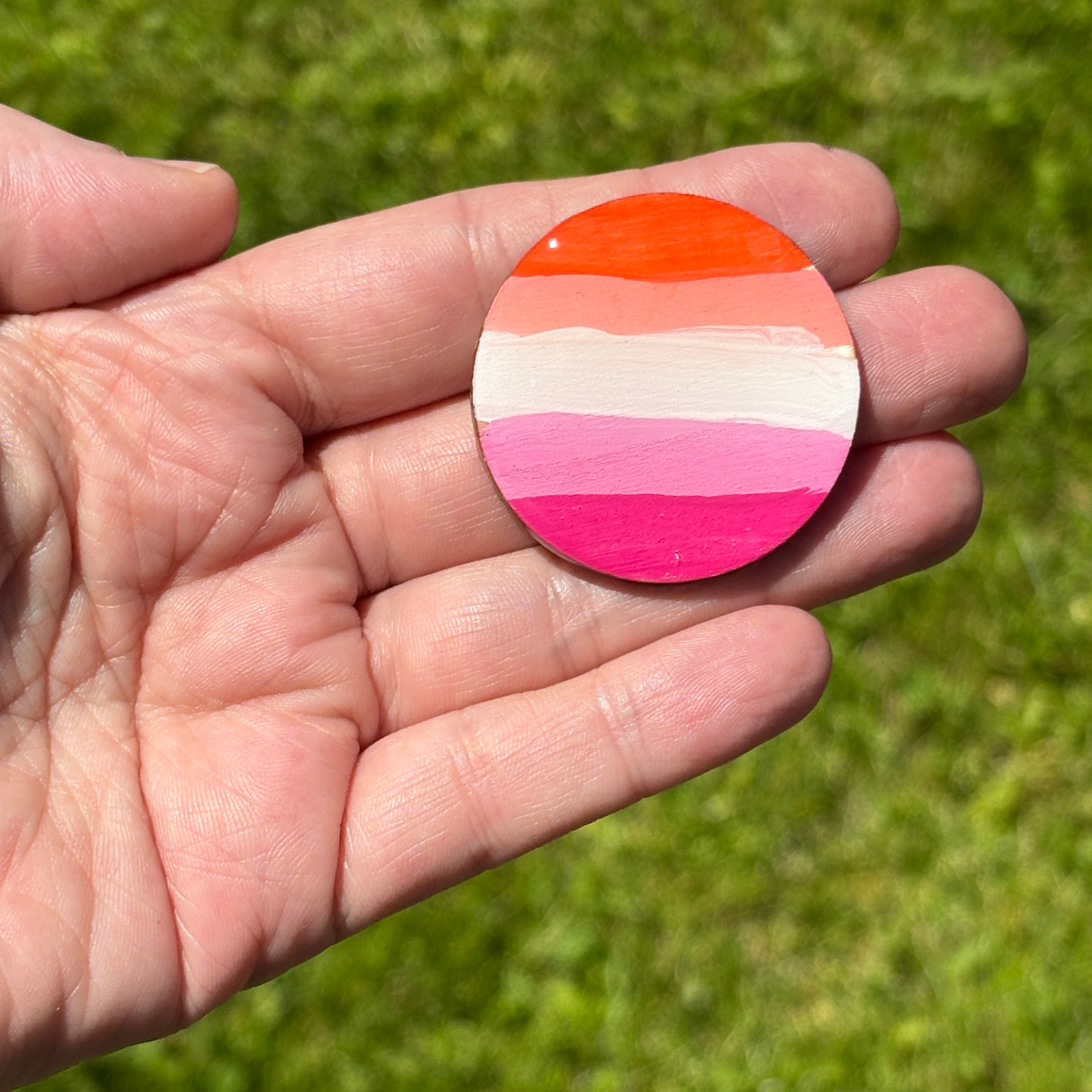 Hand holding a round magnet with hand painted lesbian flag pattern against a green grass background
