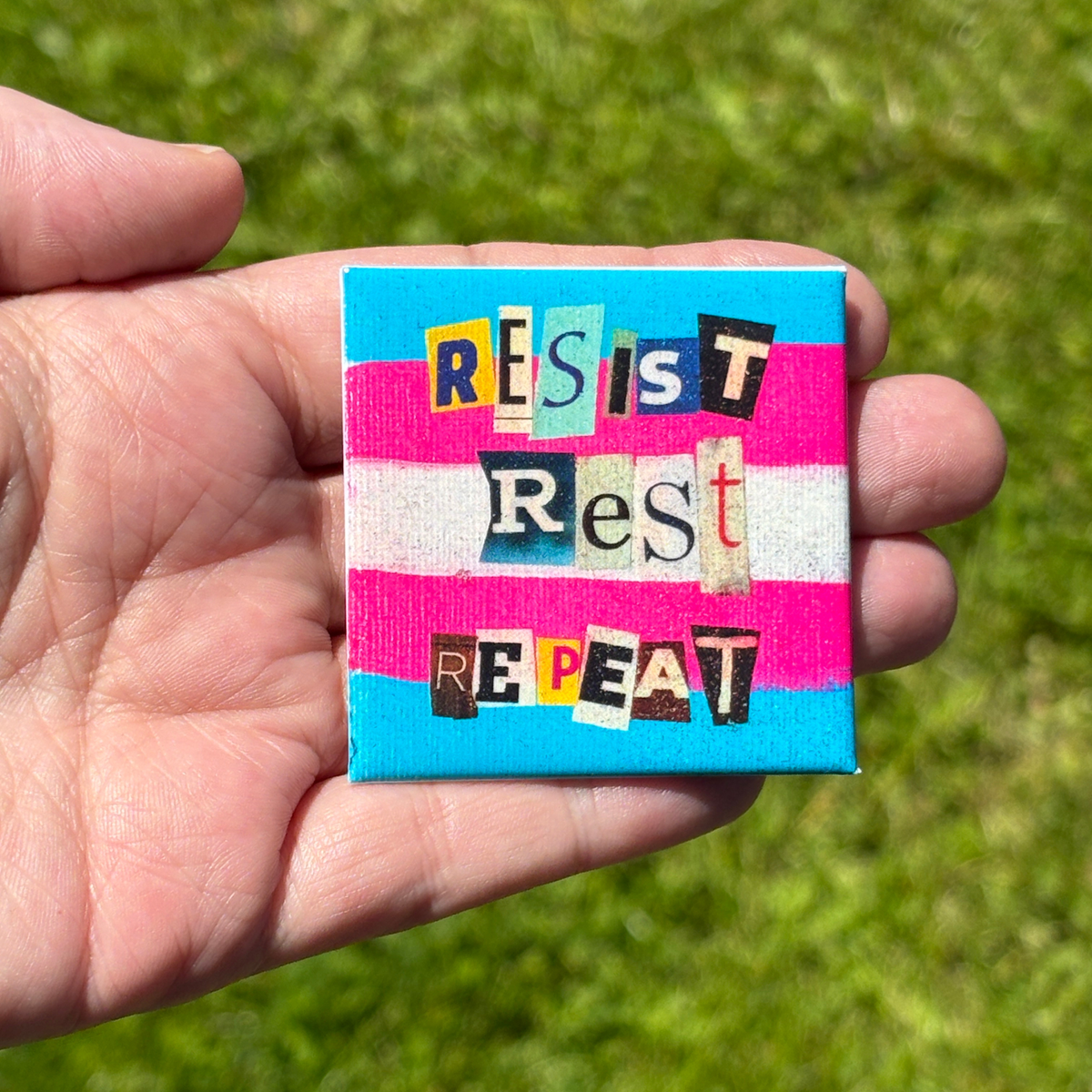 Hand holding a small square trans flag button with 'RESIST Rest REPEAT' text against a grass background