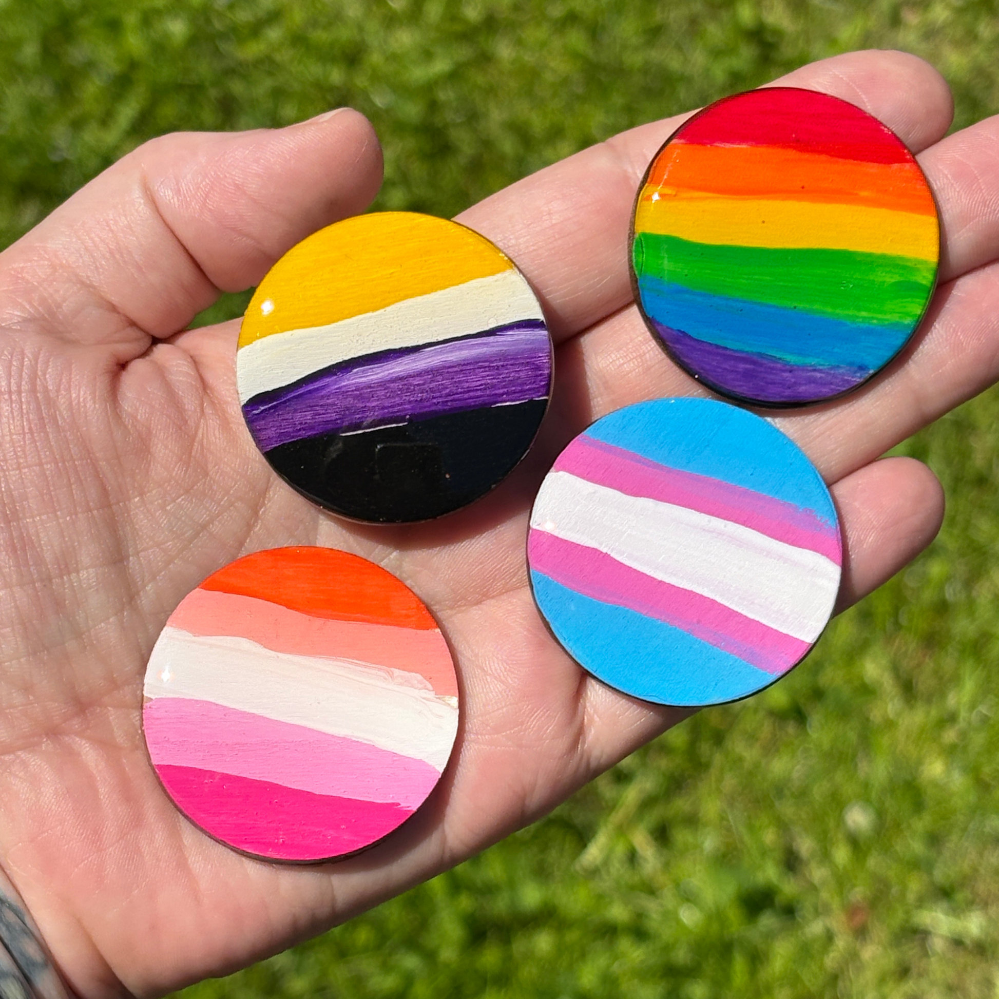Hand holding four round magnets, each with a different pride flag pattern, against a grassy background.  From left to right, they are the lesbian pride flag, the non binary pride flag, the trans pride flag, and the standard pride flag. 