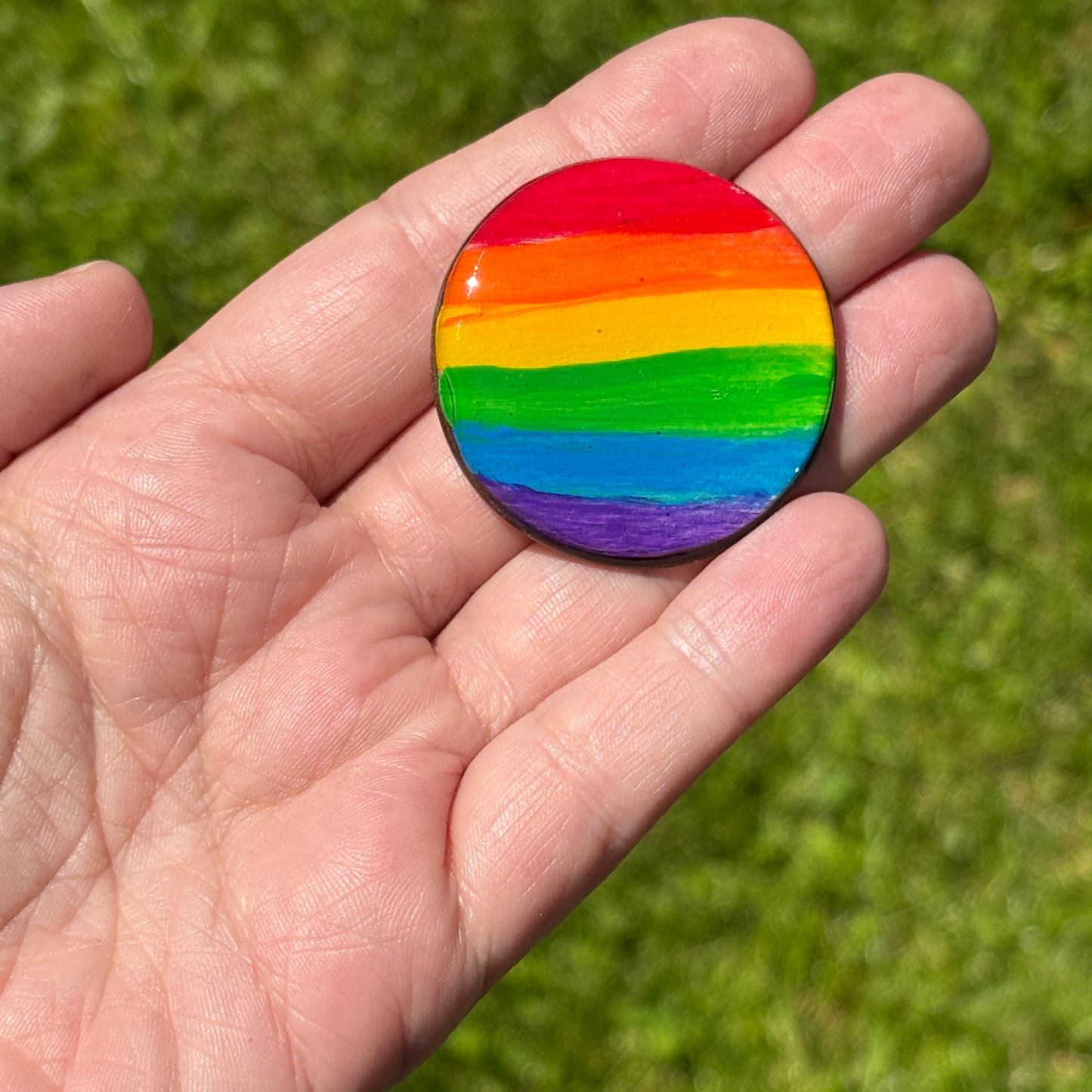 Hand holding a round magnet with hand painted pride flag pattern against a green grass background