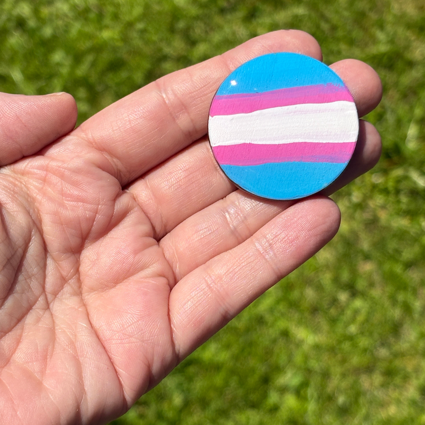 Hand holding a round magnet with hand painted trans flag pattern against a green grass background