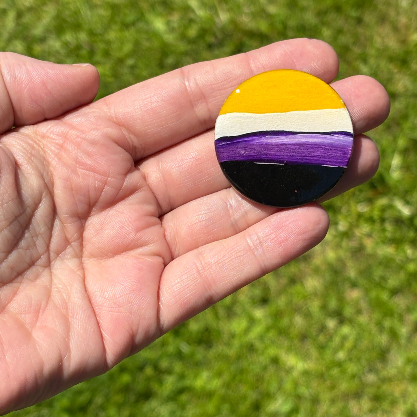 Hand holding a round magnet with a hand painted non binary flag pattern against a grassy background