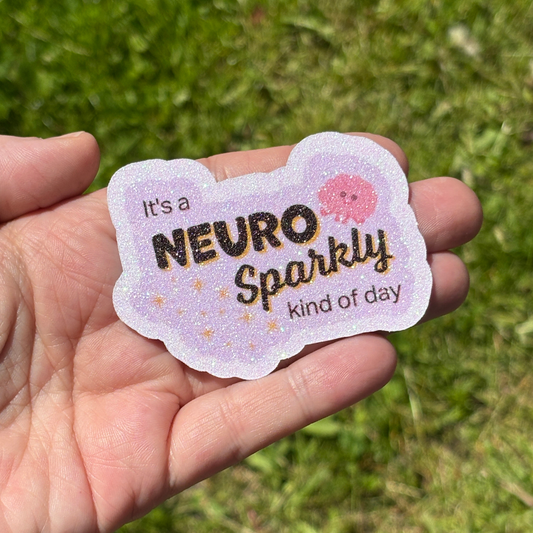Hand holding a sticker with 'It's a Neuro Sparkly kind of day' text against a grassy background