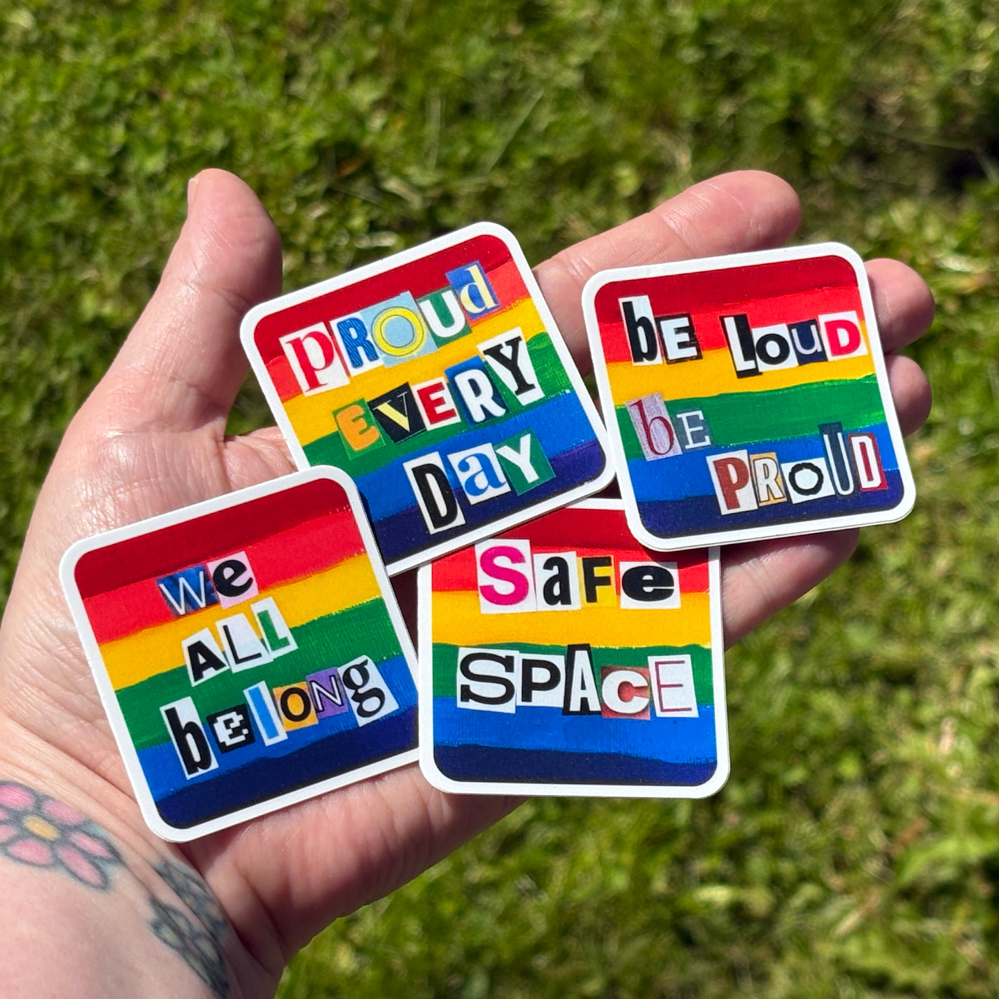 Four rainbow-colored stickers with motivational phrases held in a hand against a grassy background.