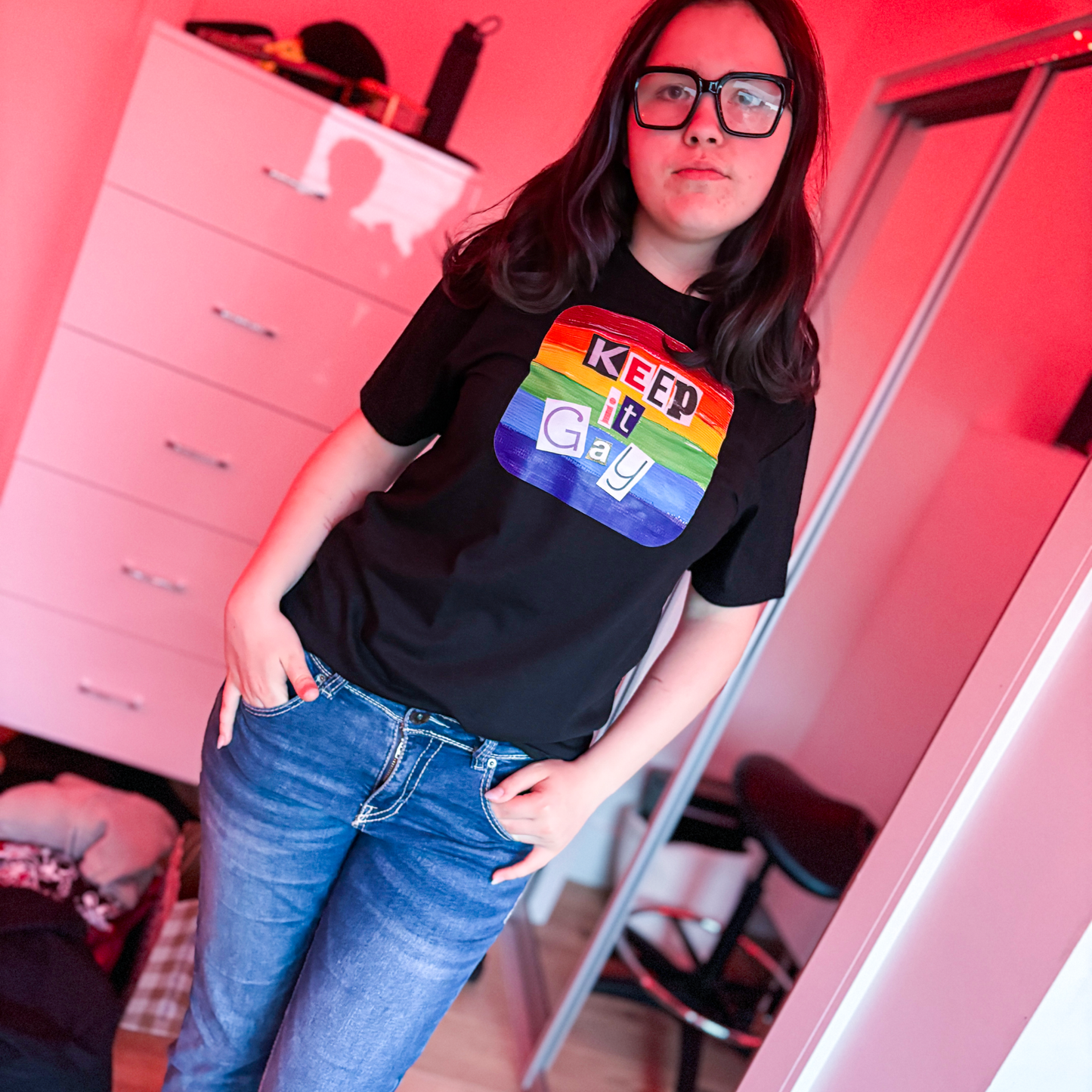 Person wearing a black t-shirt with rainbow design saying Keep it Gay in a room with red lighting.