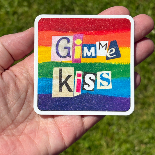 Colorful vinyl Pride sticker with 'Gimme Kiss' text held in a hand against a grass background