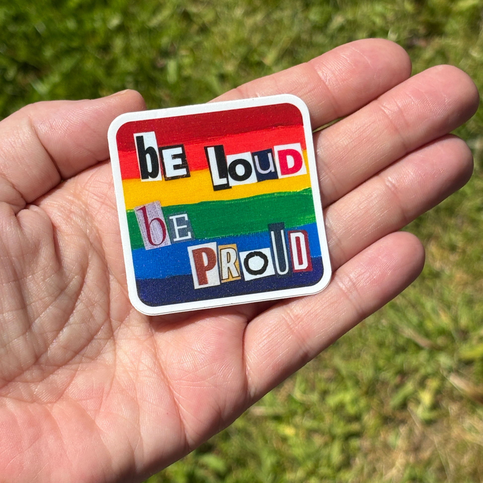 Hand holding a rainbow sticker with 'be loud be proud' text against a grassy background