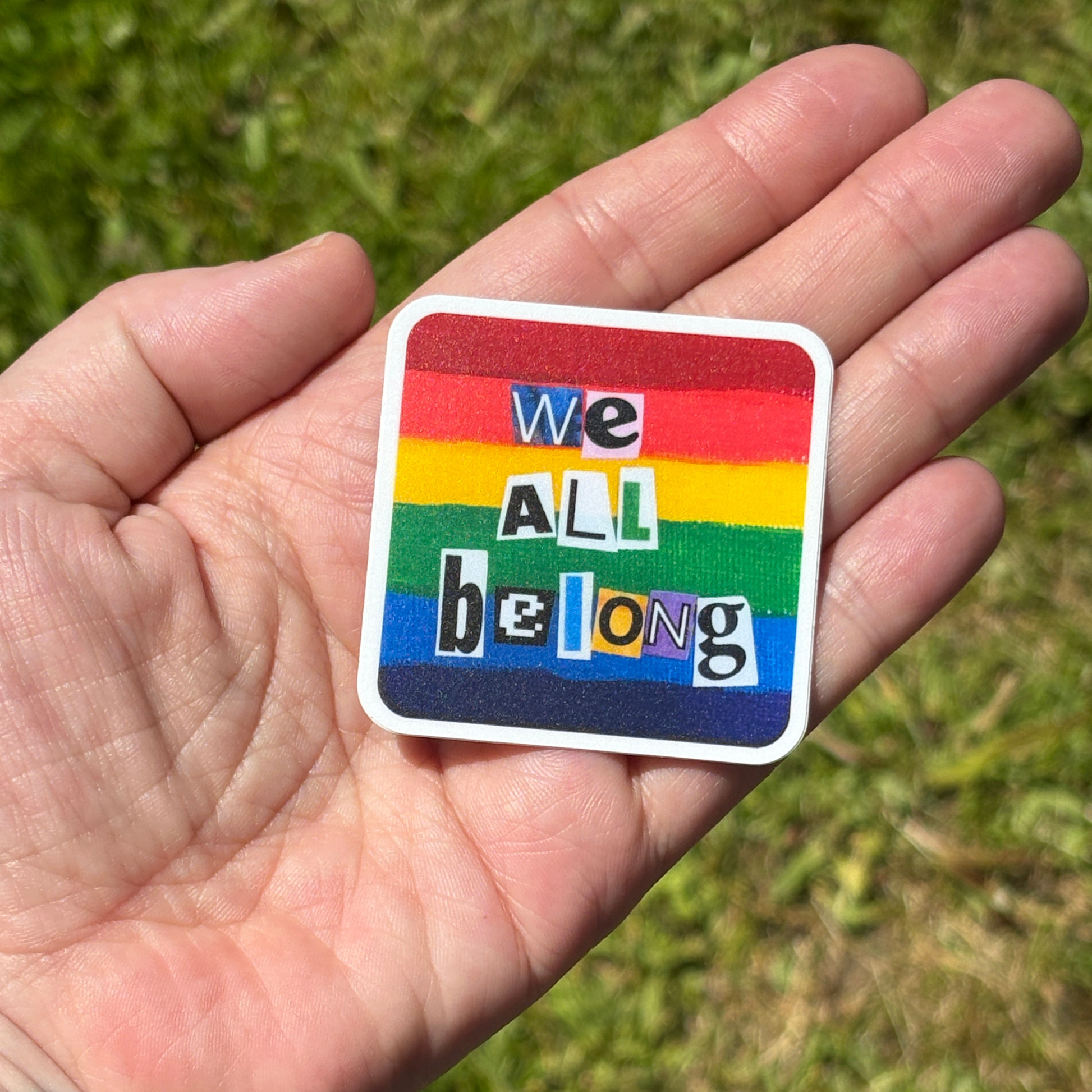 Hand holding a rainbow sticker with 'we all belong' text against a grassy background
