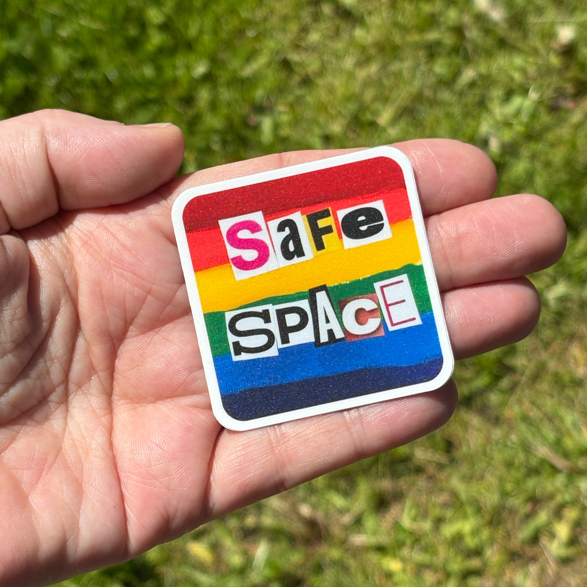 Hand holding a 'Safe Space' sticker with rainbow colors against a grassy background