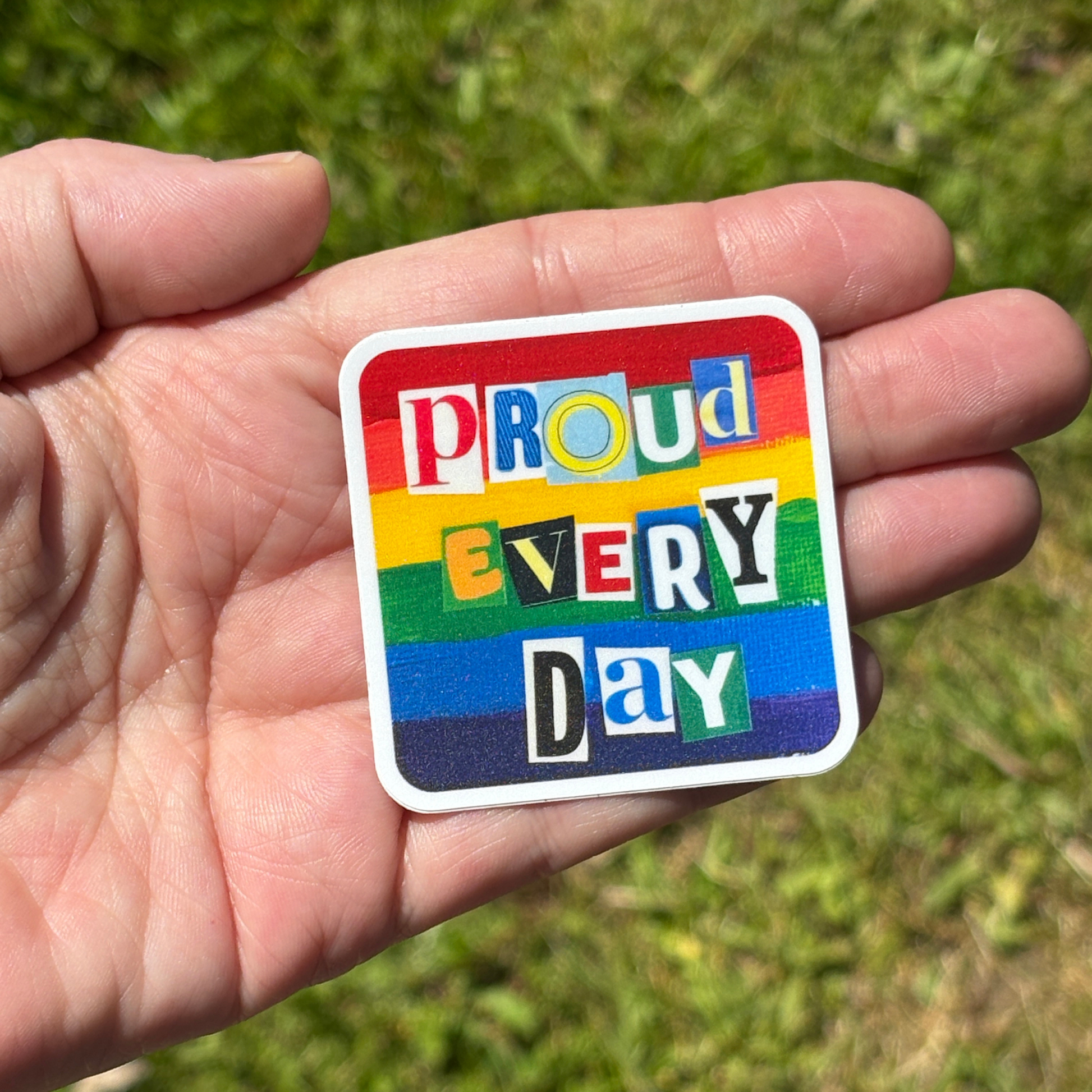 Hand holding a rainbow sticker with 'Proud Every Day' text against a grassy background