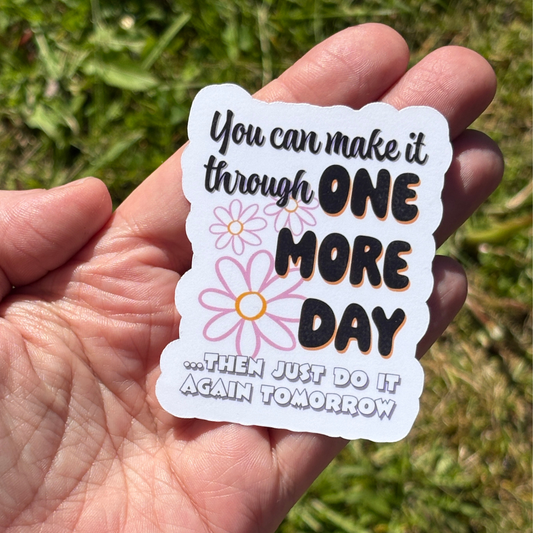 Hand holding a motivational sticker with text and flowers against a grassy background