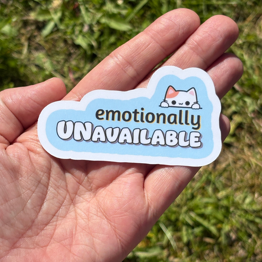 Hand holding a sticker with 'emotionally UNAVAILABLE' text and a cat illustration against a grassy background.