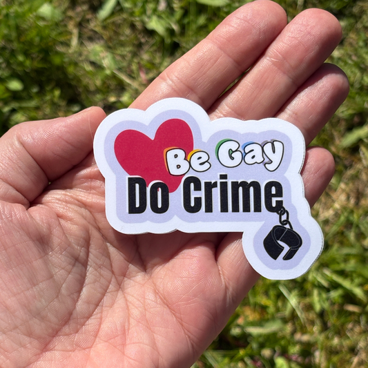 Hand holding a sticker that says "Be Gay Do Crime" and symbols against a grassy background
