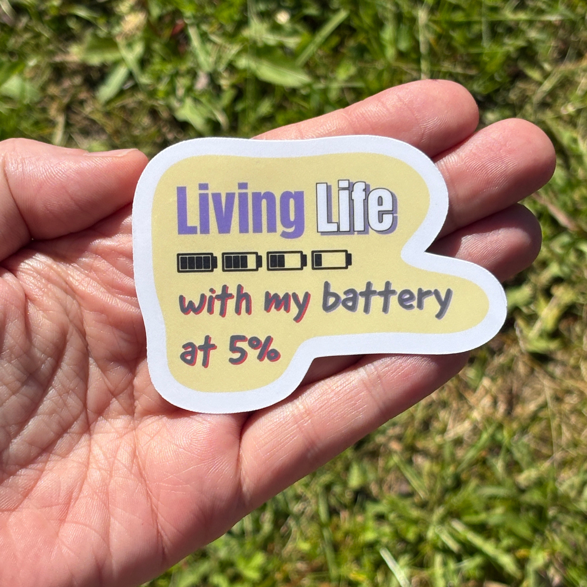 Hand holding a sticker with text 'Living Life with my battery at 5%' against a grassy background