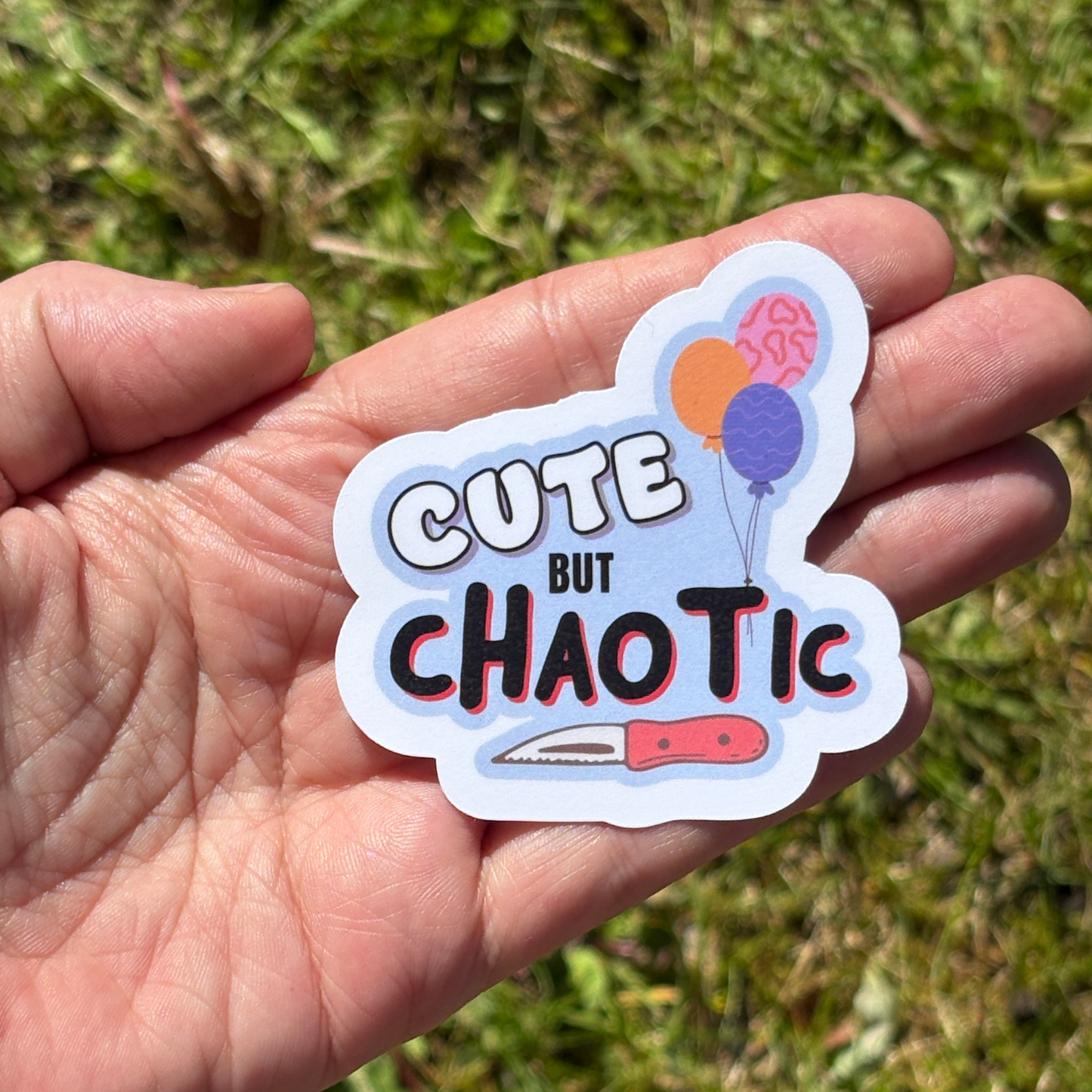 Hand holding a sticker with 'Cute But Chaotic' text and graphics against a grassy background