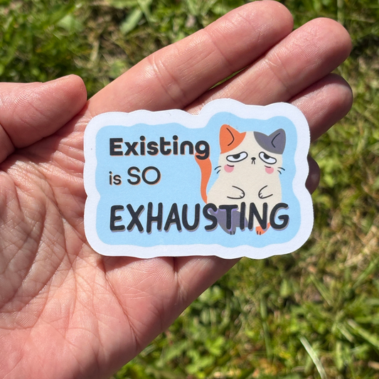 Hand holding a sticker with a cat illustration and text 'Existing is SO EXHAUSTING' against a grassy background.
