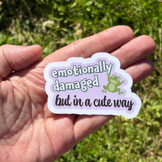 Hand holding a sticker with text 'emotionally damaged but in a cute way' and a frog graphic against a grassy background.
