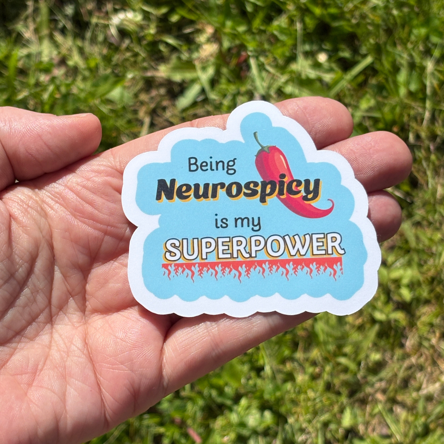 Hand holding a sticker with 'Being Neurospicy is my SUPERPOWER' text against a grassy background