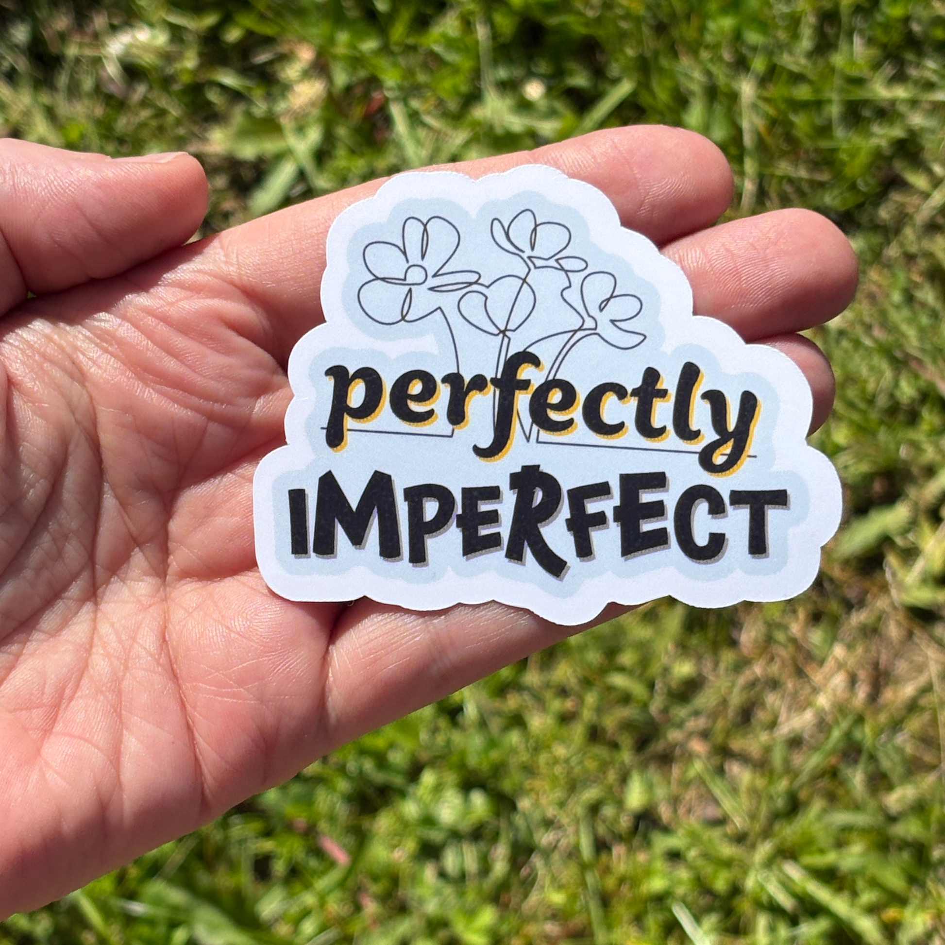 Hand holding a 'perfectly imperfect' sticker with floral design against a grassy background