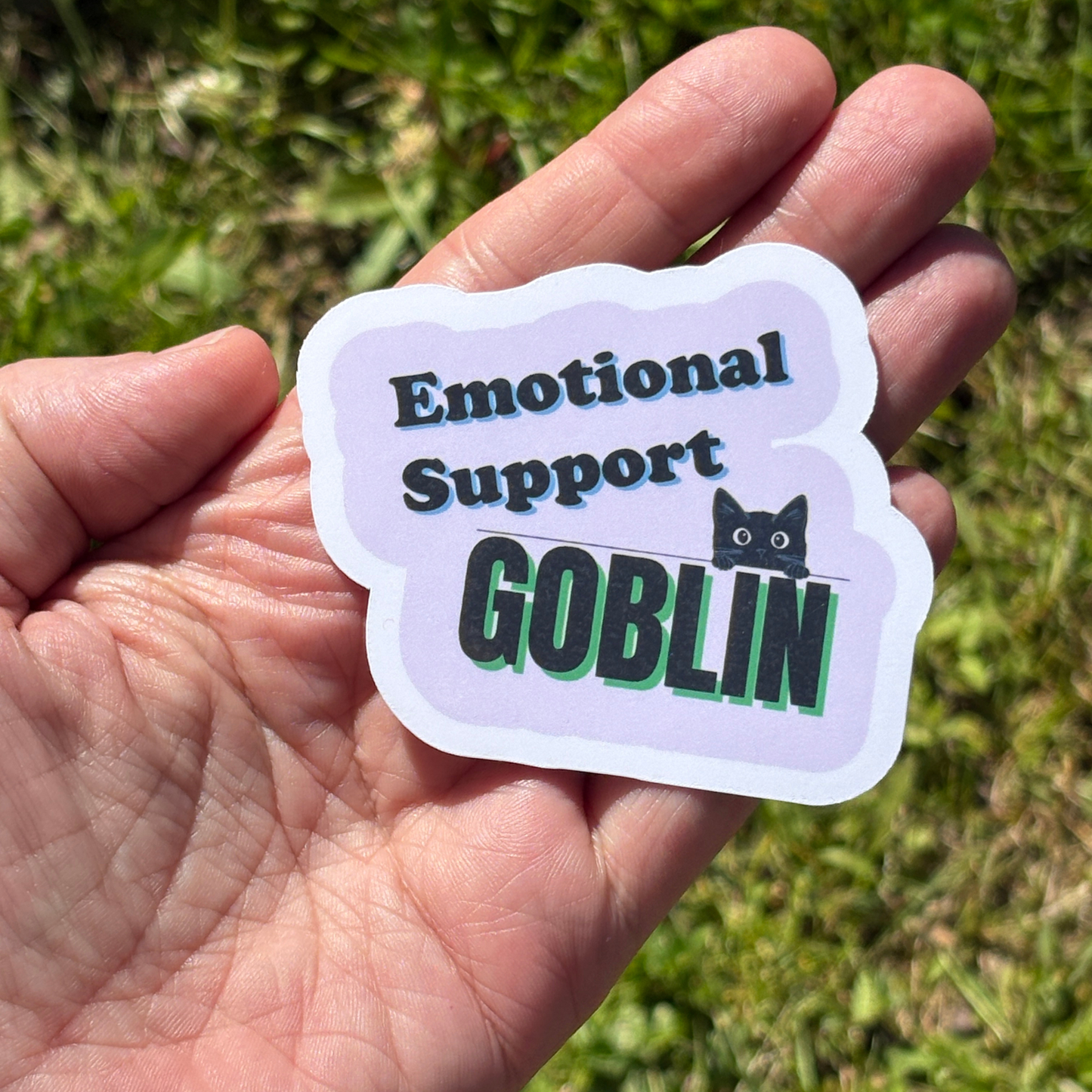 Hand holding a sticker with 'Emotional Support Goblin' text and a cat icon against a grassy background.