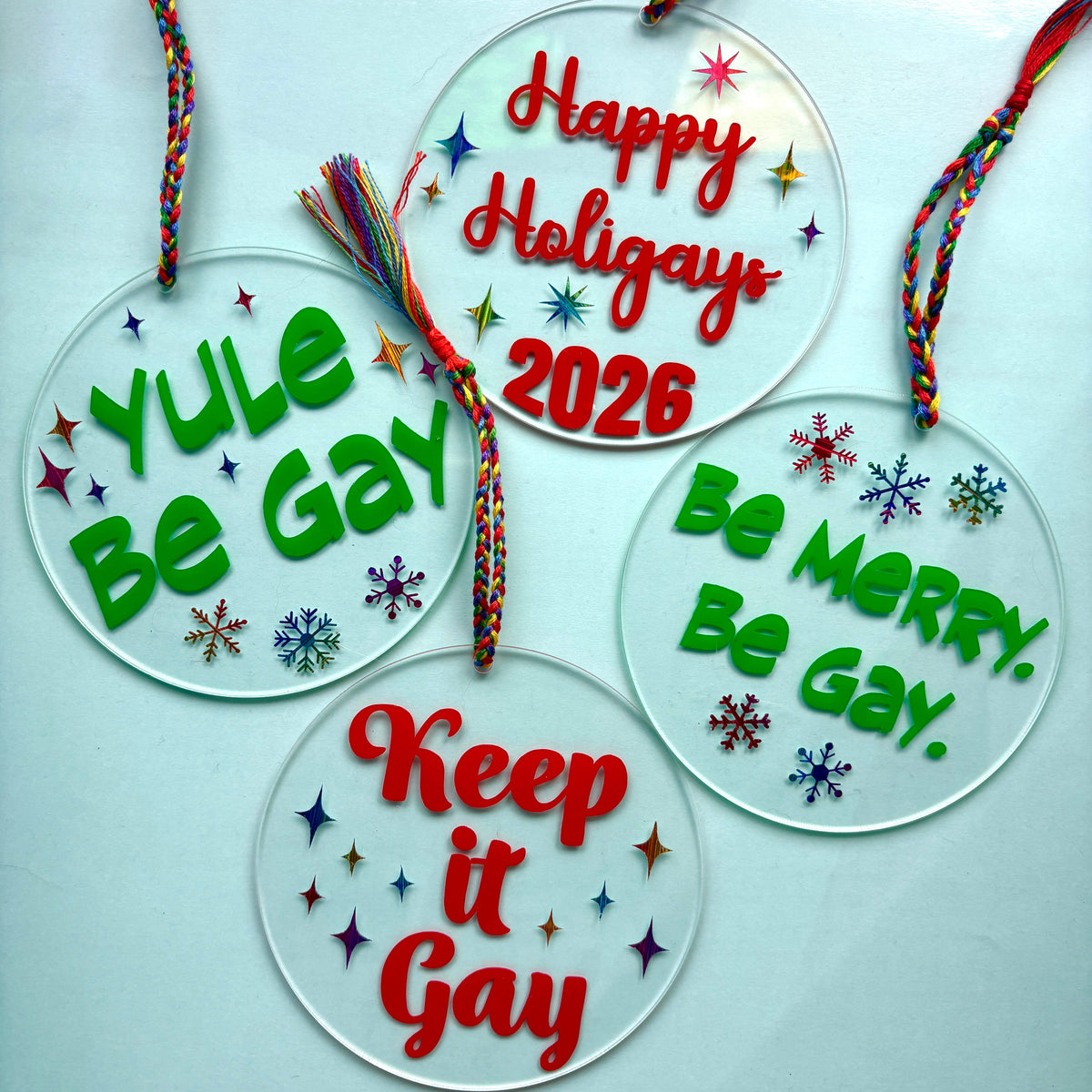 Four round clear acrylic holiday ornaments with colorful braided hanging cords, decorated with red and green text and small multicolored snowflakes. The ornaments read: 'Yule Be Gay,' 'Happy Holidays 2026,' 'Be Merry, Be Gay,' and 'Keep it Gay,' displayed on a pale blue background.