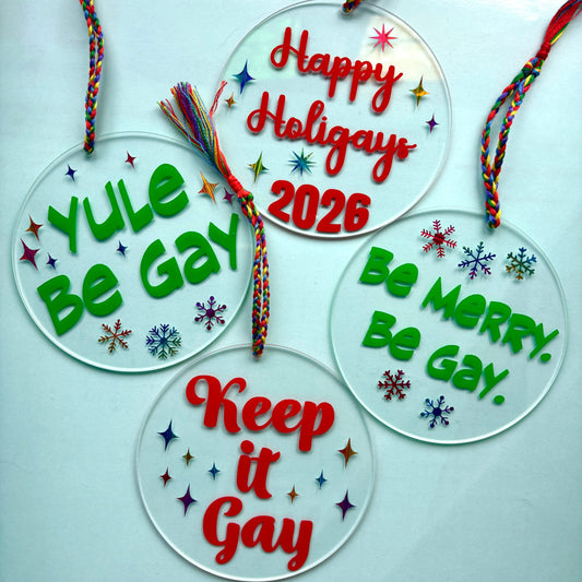 Four round clear acrylic holiday ornaments with colorful braided hanging cords, decorated with red and green text and small multicolored snowflakes. The ornaments read: 'Yule Be Gay,' 'Happy Holidays 2026,' 'Be Merry, Be Gay,' and 'Keep it Gay,' displayed on a pale blue background.