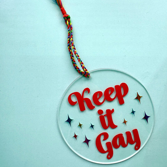 round acrylic Keep it Gay Christmas ornament hanging from a hand braided rainbow hanger.