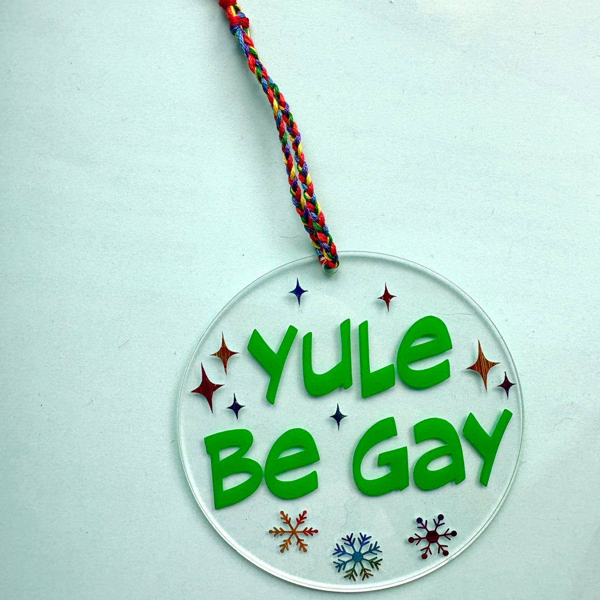 A round clear acrylic hanging ornament with the words 'YULE Be Gay' in bold green letters, decorated with small colorful star and snowflake cutouts, and suspended by a braided multicolor tassel on a pale blue background.