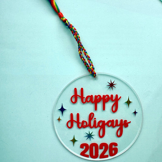 A round clear acrylic holiday ornament with 'Happy Holigays 2026' written in red script and numbers, decorated with small colorful star and diamond shapes, and hanging from a multicolored braided cord with a tassel, shown on a light blue background.