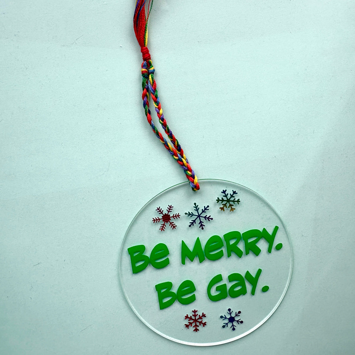 A clear circular acrylic hanging ornament with the phrase 'Be MERRY. Be GAY.