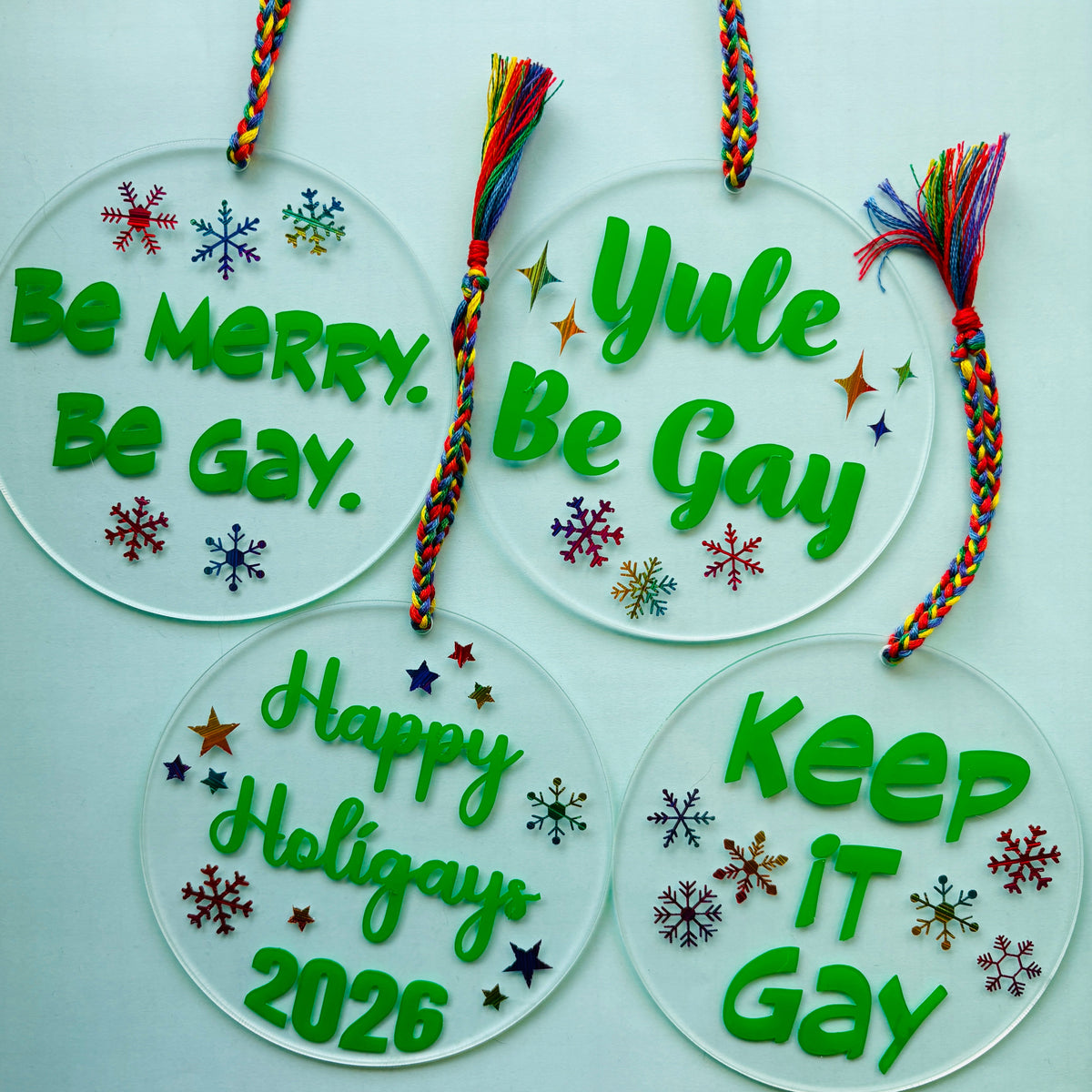Four round clear acrylic holiday ornaments with colorful braided hanging cords and tassels, each printed with green festive text and small multicolored snowflake and star designs. The text reads: 'Be Merry.
