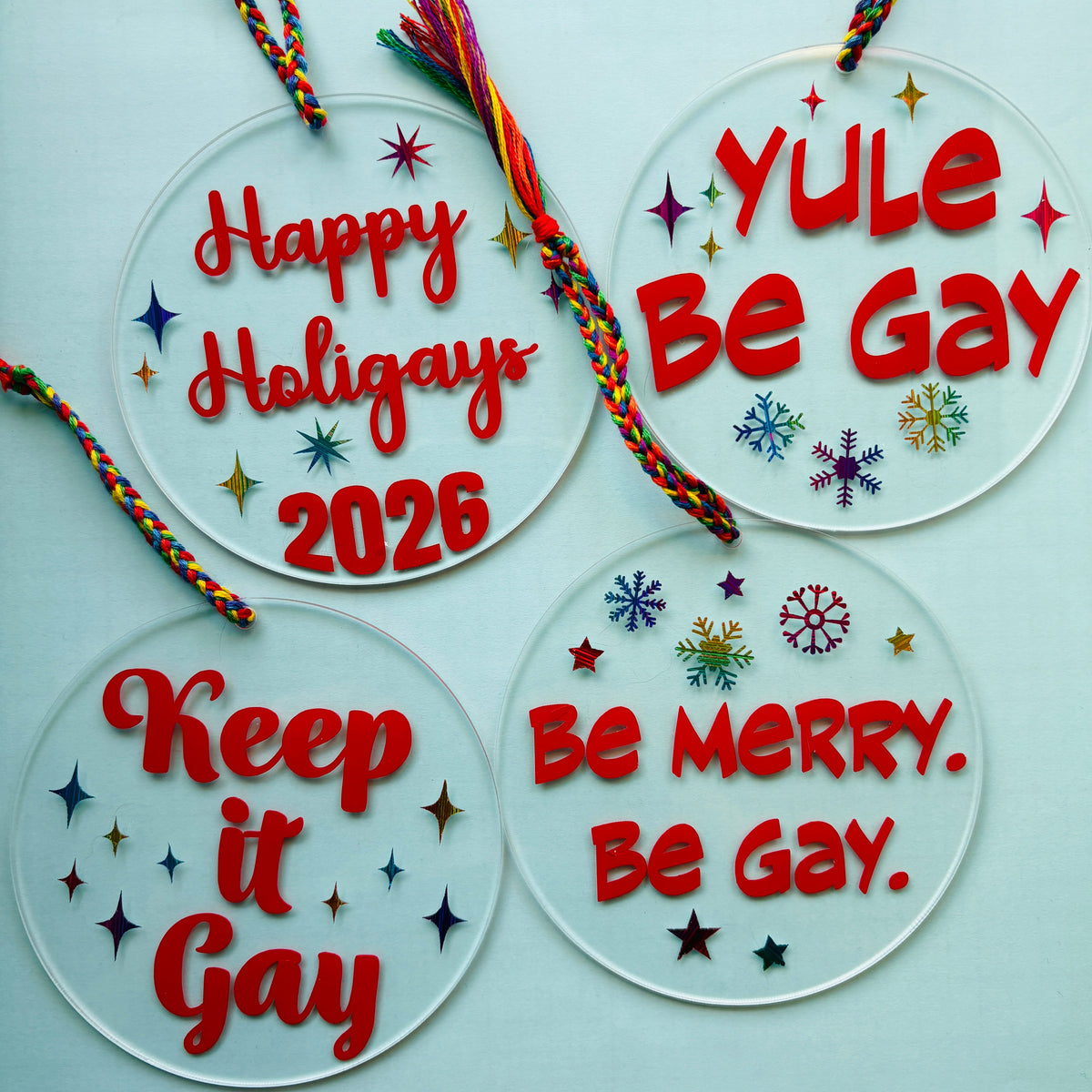Four round clear acrylic holiday ornaments with colorful braided hanging cords, decorated with red text and small multicolored stars and snowflake designs, displayed on a pale blue background.