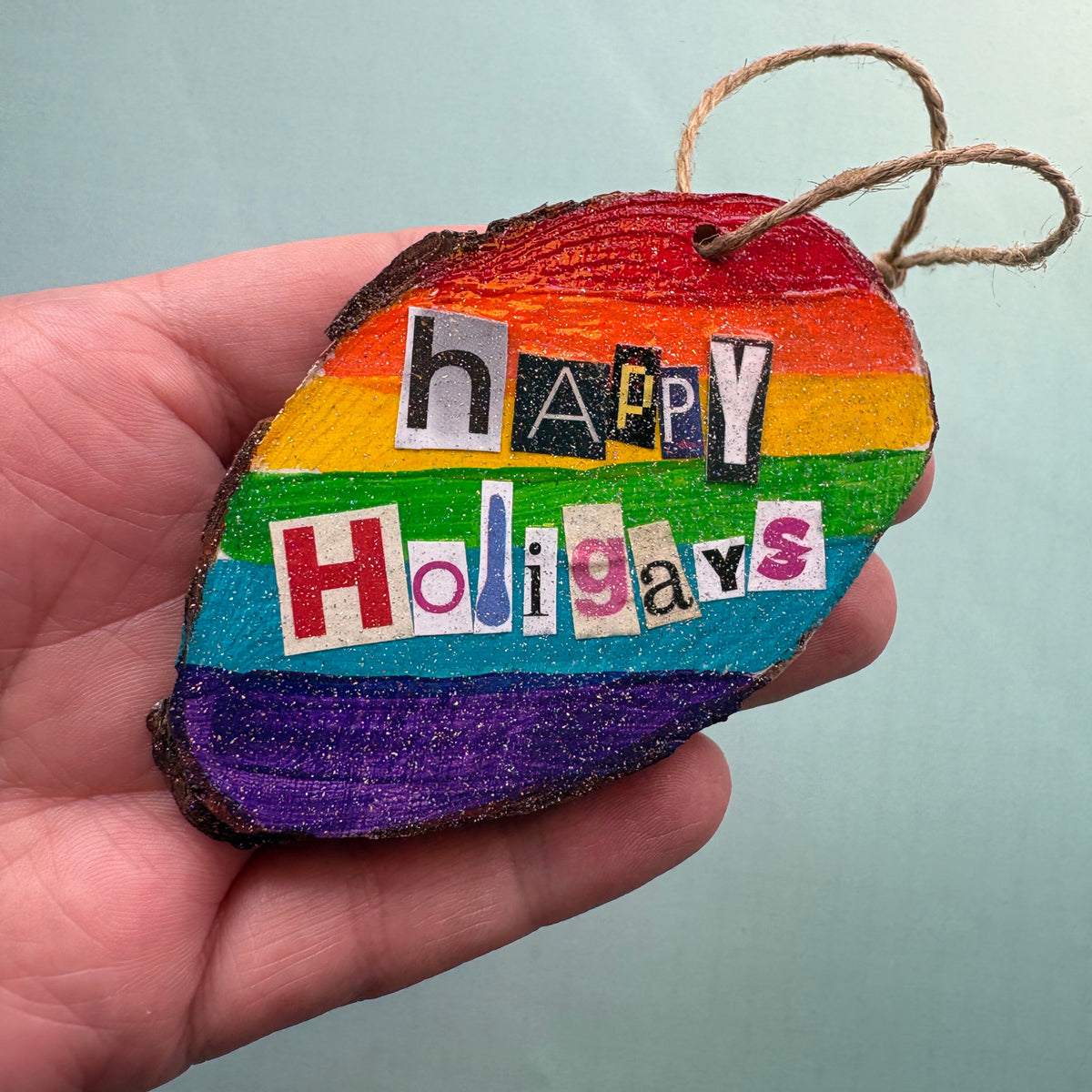 wooden christmas decoration hand painted with a rainbow background. The phrase Happy Holigays is spelled out in collage style lettering. 