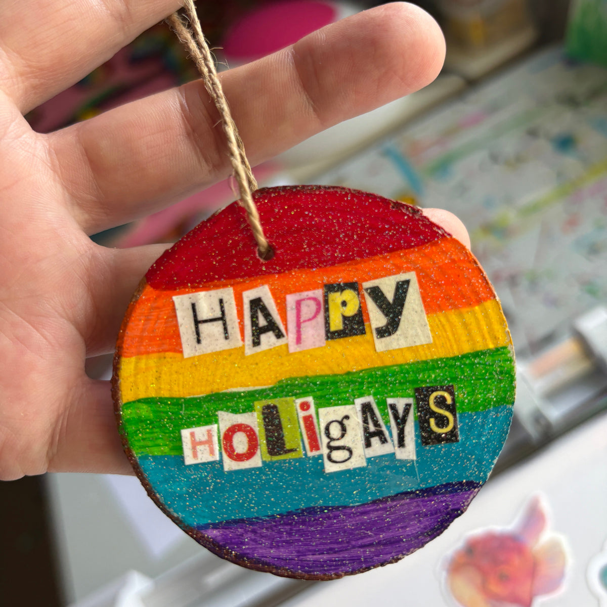 wooden christmas tree decoration, hand painted with a rainbow background. The phrase Happy Holigays is spelled out in collage style lettering
