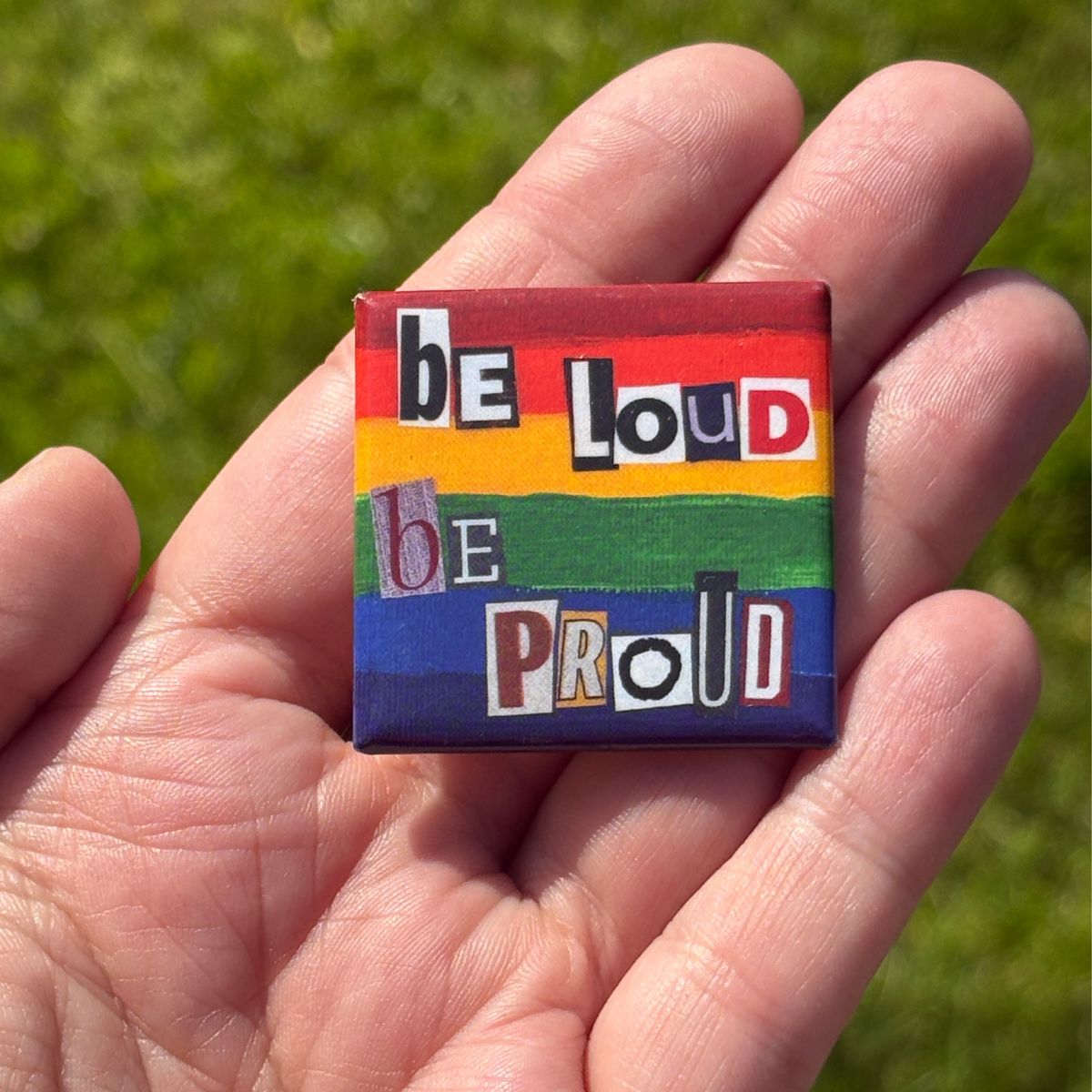a 1.5" pride flag button with 'be loud be proud' text held in a hand against a grassy background