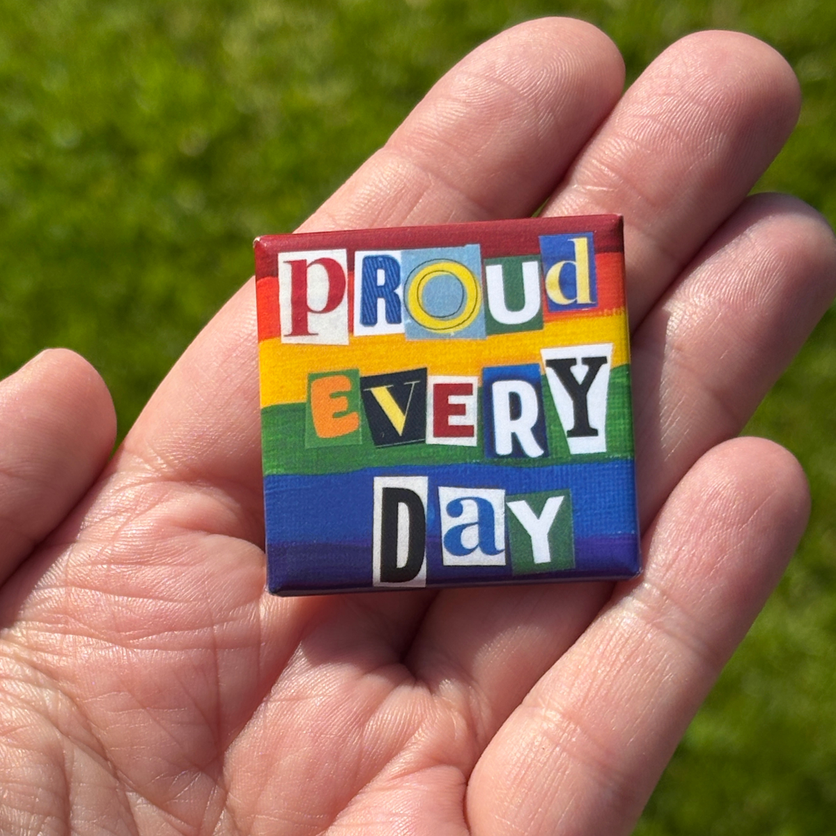 a 1.5" pride flag button with 'Proud Every Day' text held in a hand against a grassy background