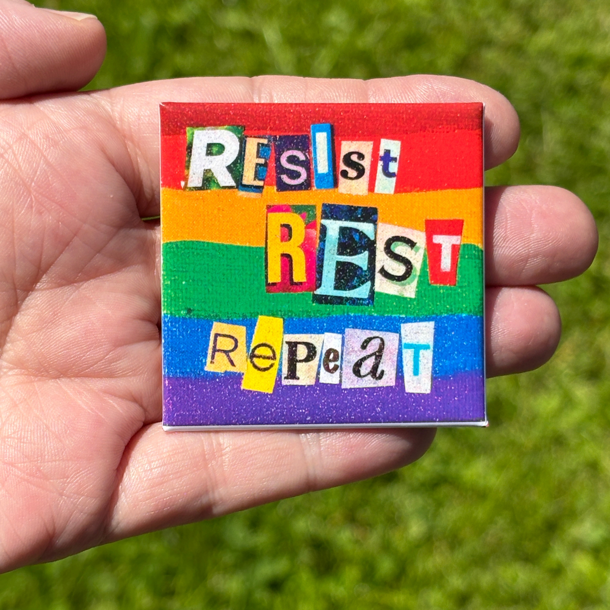 Colorful square pride flag button with 'Resist Rest Repeat' text held in a hand against a grass background