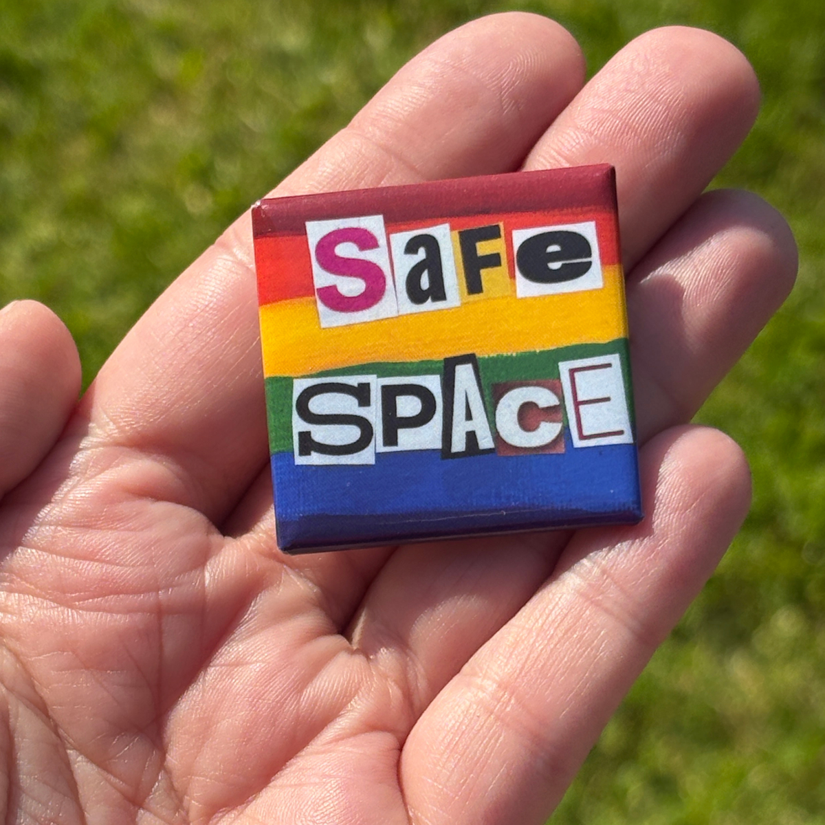 a 1.5" pride flag button with 'safe space' text held in a hand against a grassy background