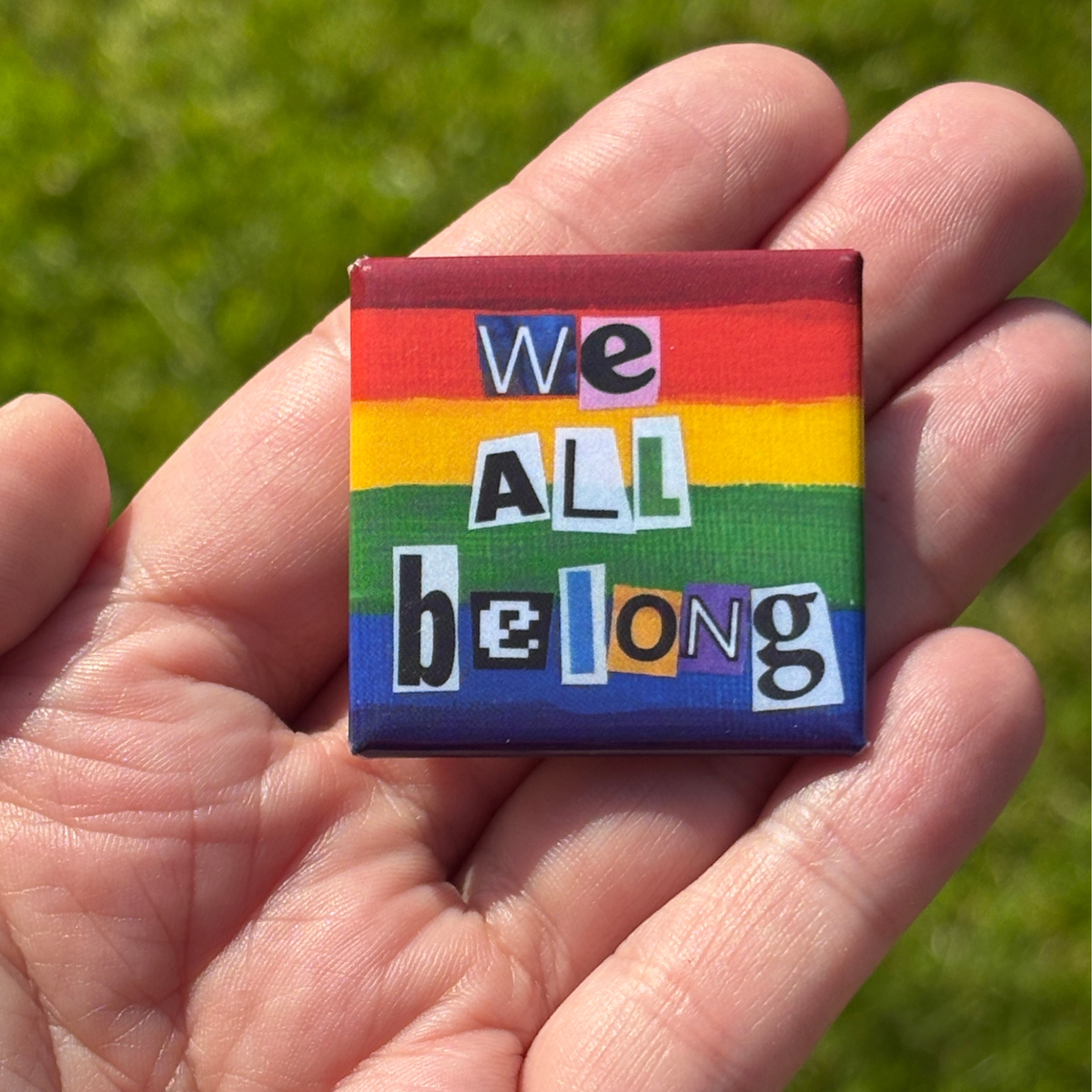 a 1.5" pride flag button with 'We All Belong' text held in a hand against a grassy background