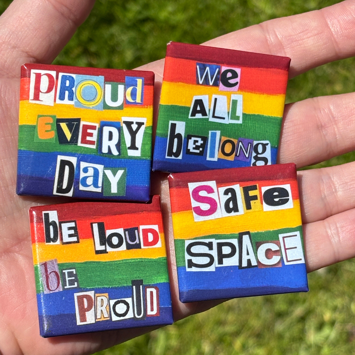 Four 1.5" pride flag buttons, each with a statement in collage letters, held against a grassy background. The statements are "Proud Every Day", "We all Belong", "Be Loud Be Proud", and "Safe Space"