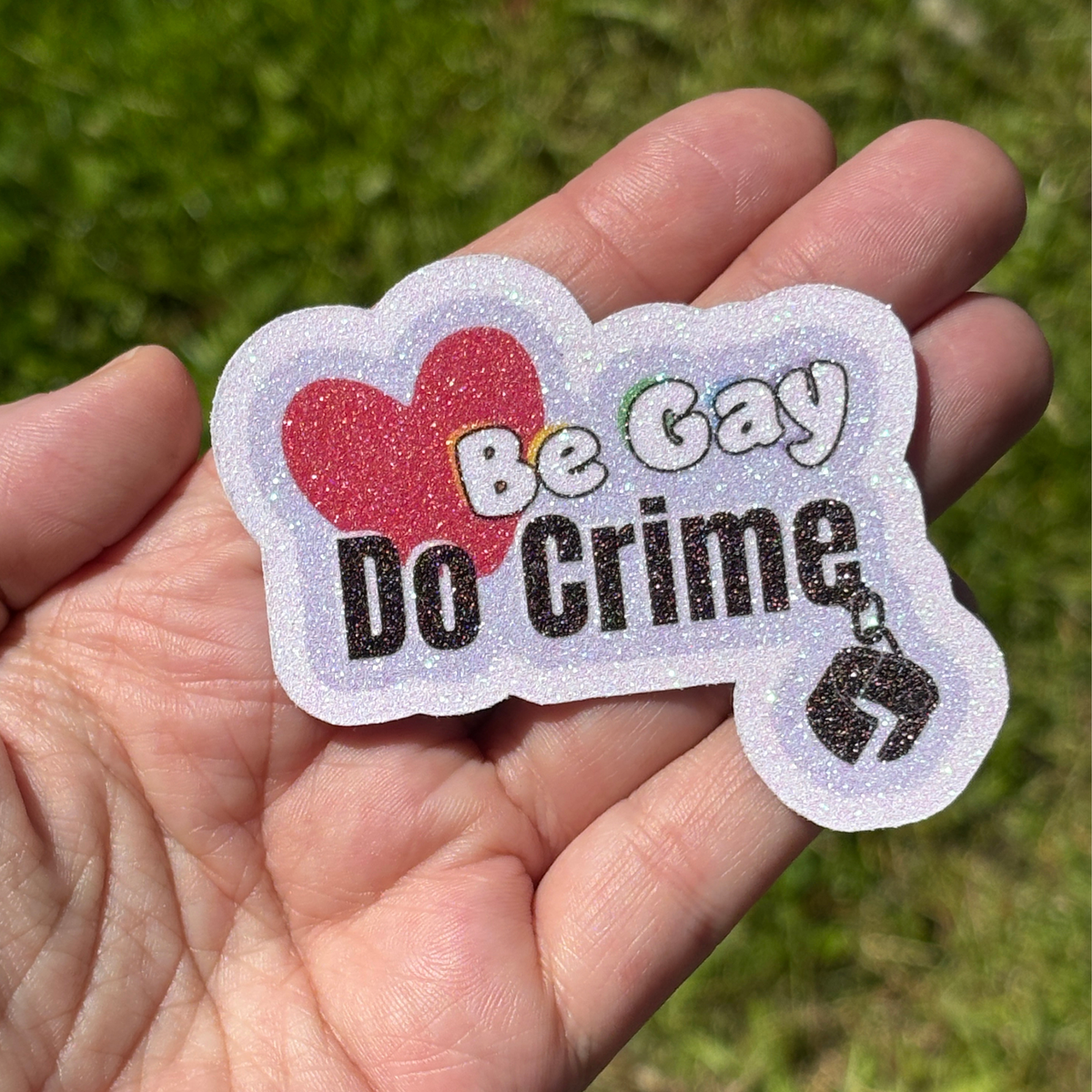 Sticker with 'Be Gay Do Crime' text held in a hand against a grass background