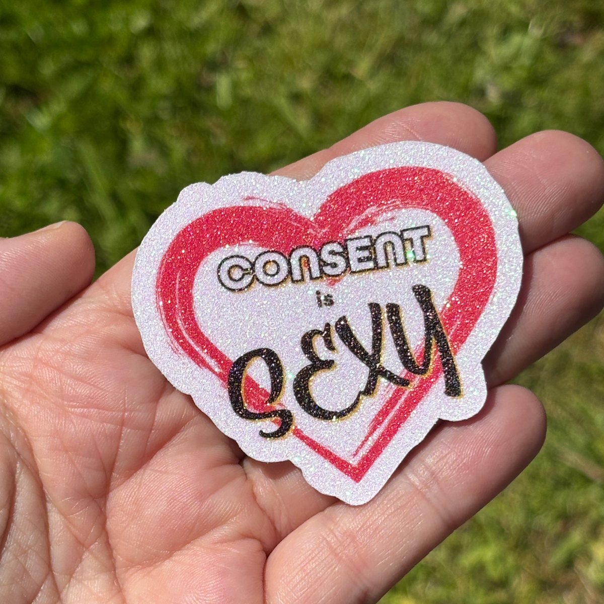 Heart-shaped sticker with 'CONSENT is SEXY' text held in a hand against a grassy background