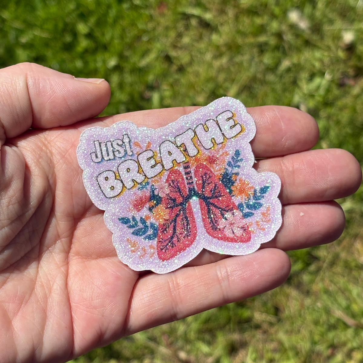Hand holding a colorful sticker with 'Just Breathe' text and a butterfly design against a grassy background.
