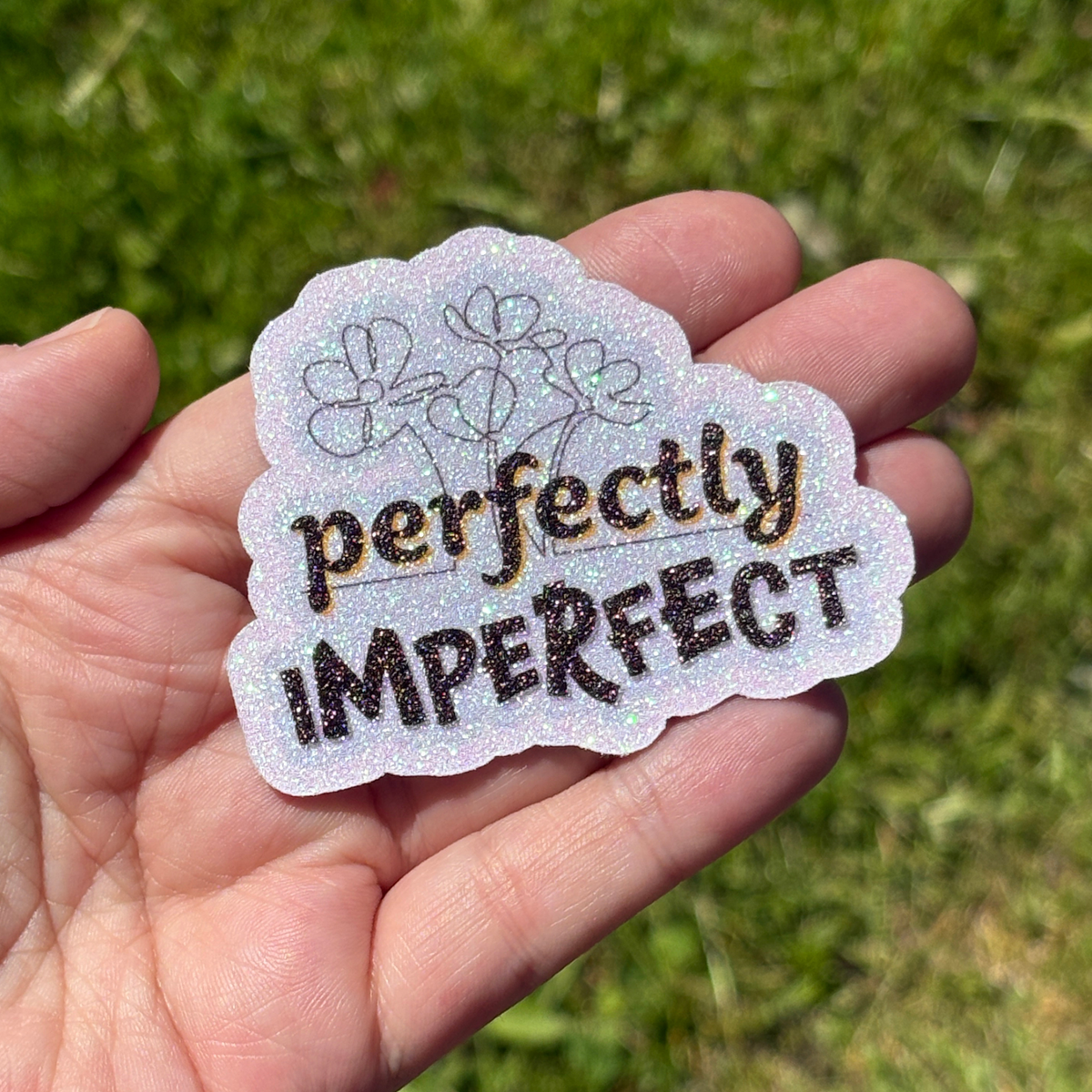 Hand holding a glittery sticker with 'perfectly imperfect' text against a grass background