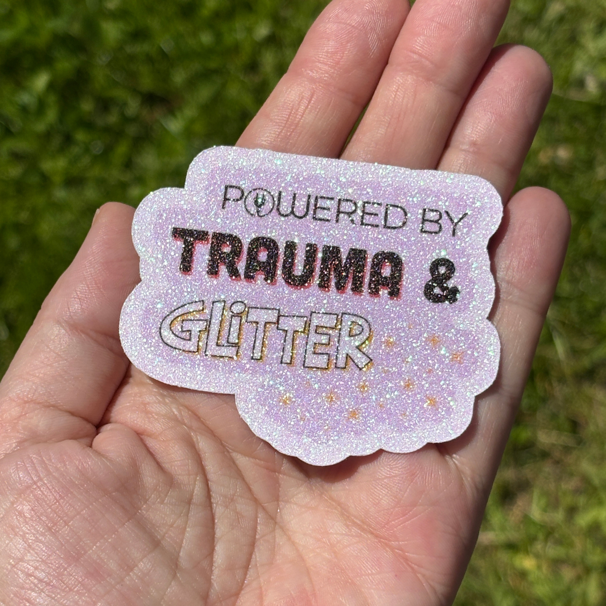 Hand holding a glittery sticker with text 'Powered by Trauma & Glitter' against a grass background