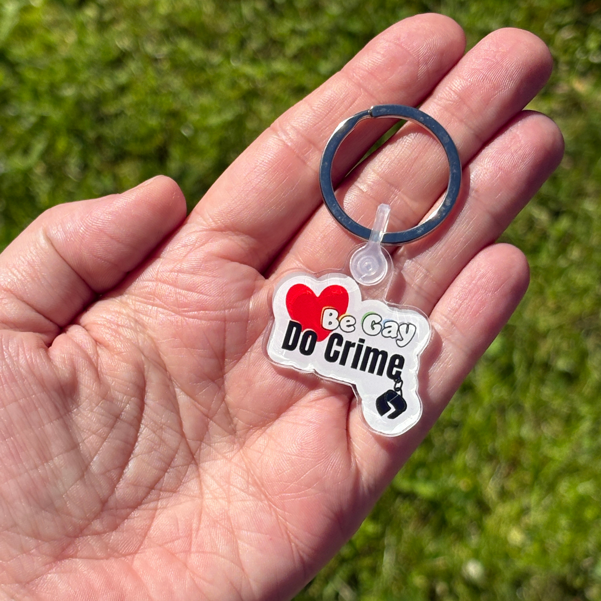 Keychain with 'Be Gay, Do Crime' text held in a hand against a grassy background