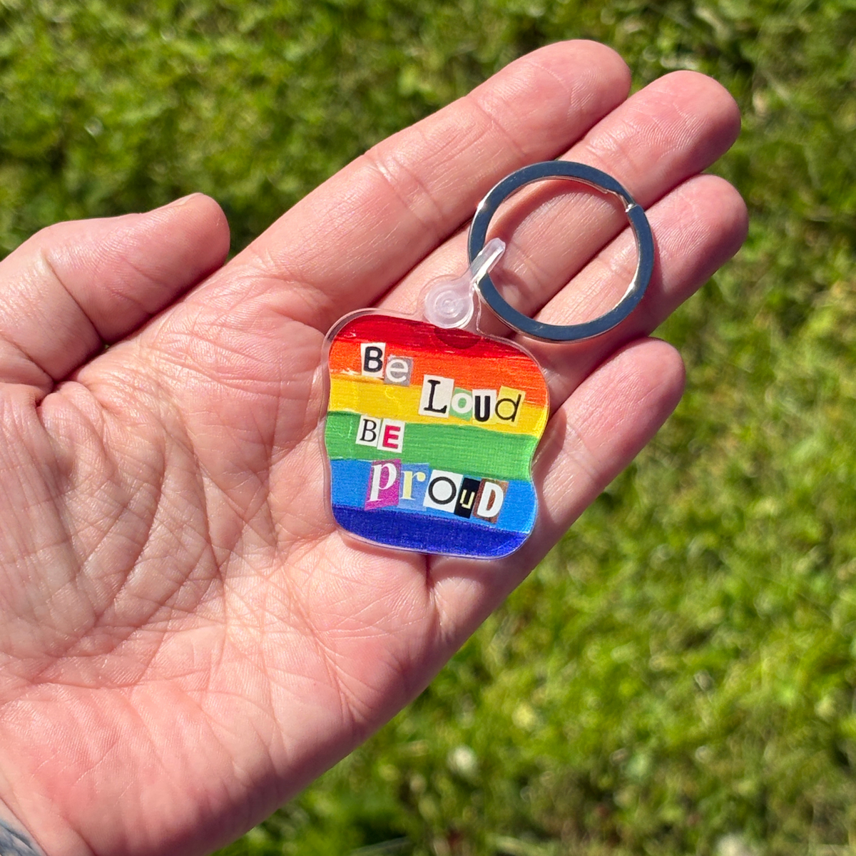 Hand holding a rainbow-colored keychain with 'Be Loud Be Proud' text against a grassy background