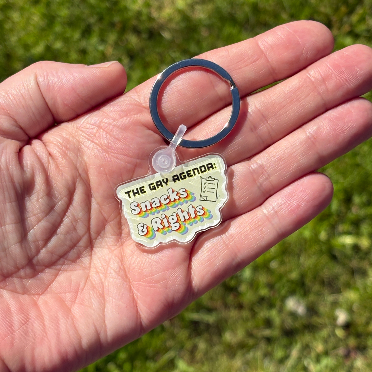 Keychain with 'The Gay Agenda: Snacks & Rights' text held in a hand against a grassy background