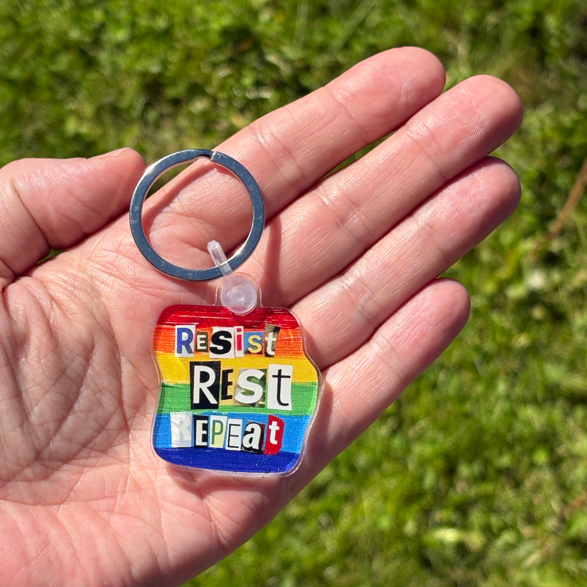 Hand holding a rainbow keychain with 'Resist Rest Repeat' text against a grassy background