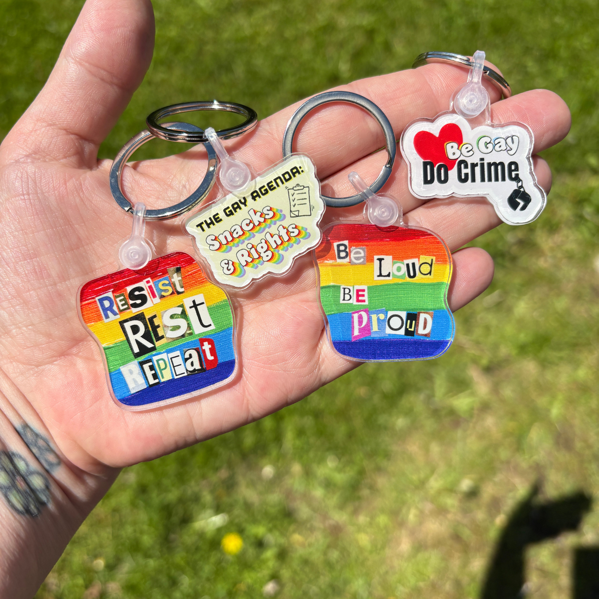 Hand holding four keychains with Pride-themed messages against a grassy background
